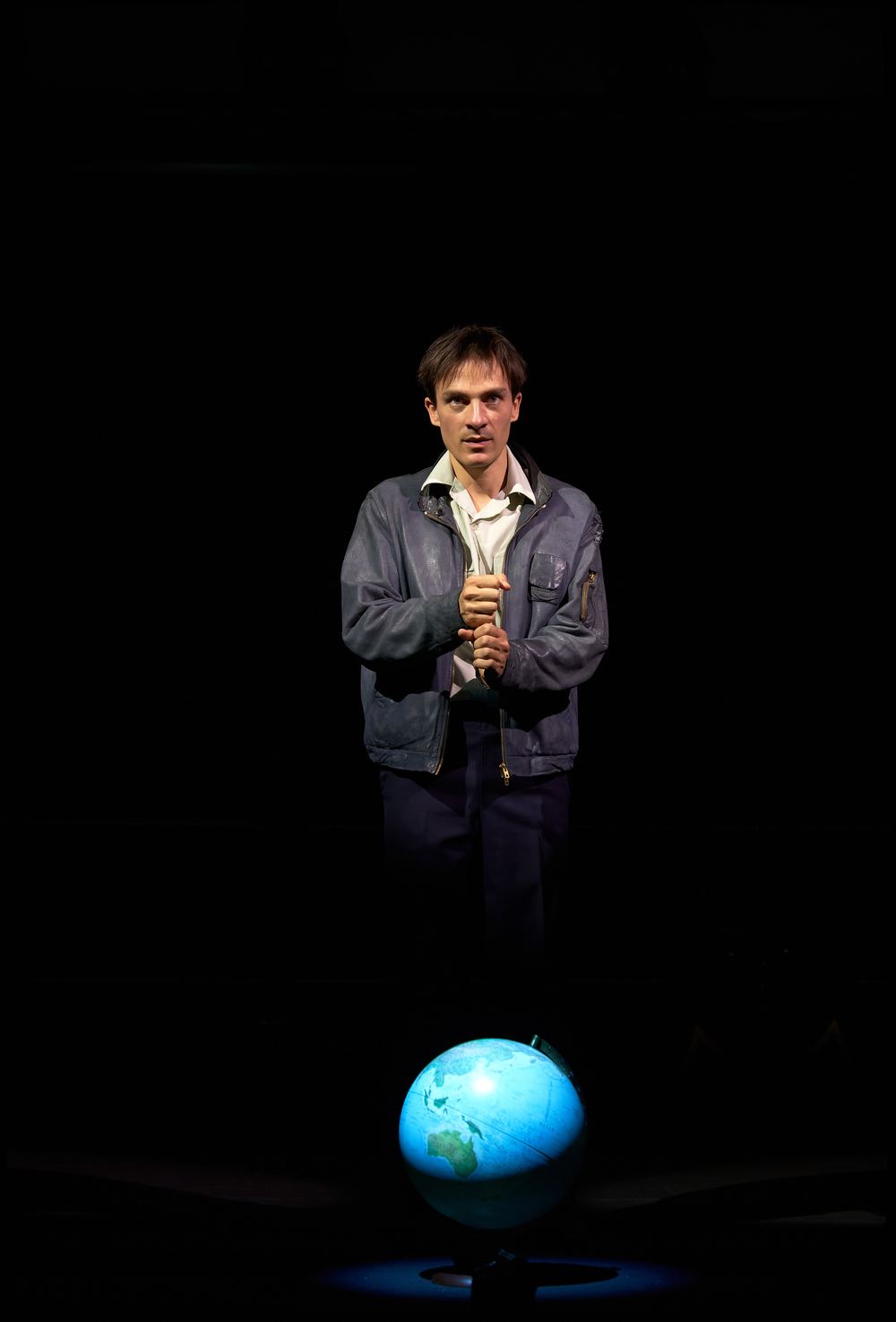 Pierre Devaux debout dans un faisceau de lumière, devant un globe terrestre, seul en scène dans Terre des Hommes